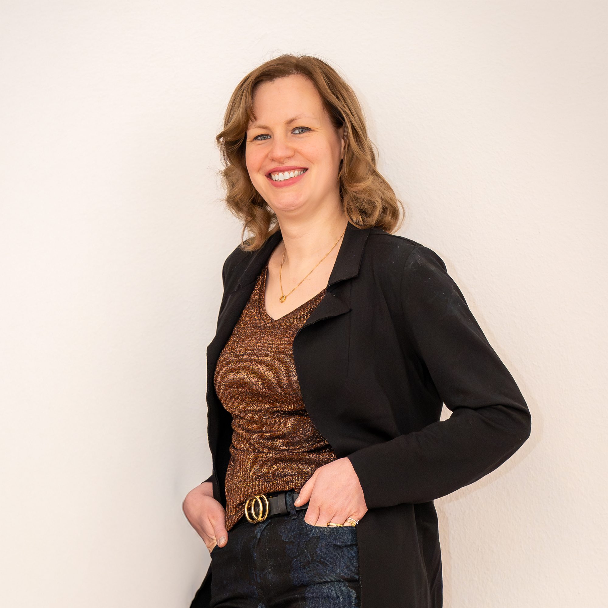 Porträt von Ines Bergel: Freundlich lächelnde Frau mit welligem Haar, in schwarzem Blazer und bronzefarbenem Glitzershirt, die Hände in den Hosentaschen.