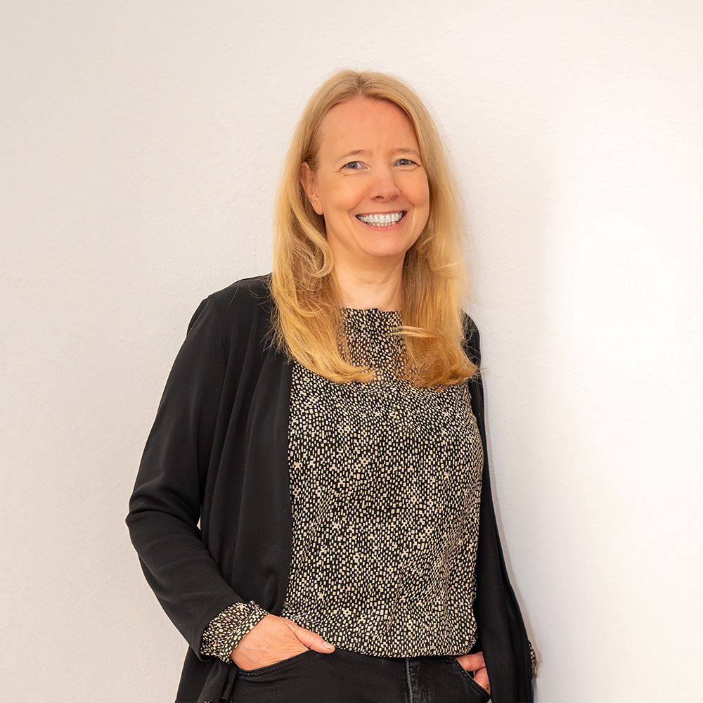 Porträt von Simone Wolf: Freundlich lachend in schwarzer Strickjacke und gemustertem Oberteil, die Hände lässig in den Hosentaschen.